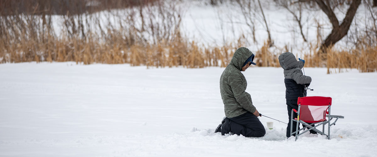 A man with a fishing rod kneels on snow-covered ice. A child stands in front of a chair adjusting their fishing pole.
