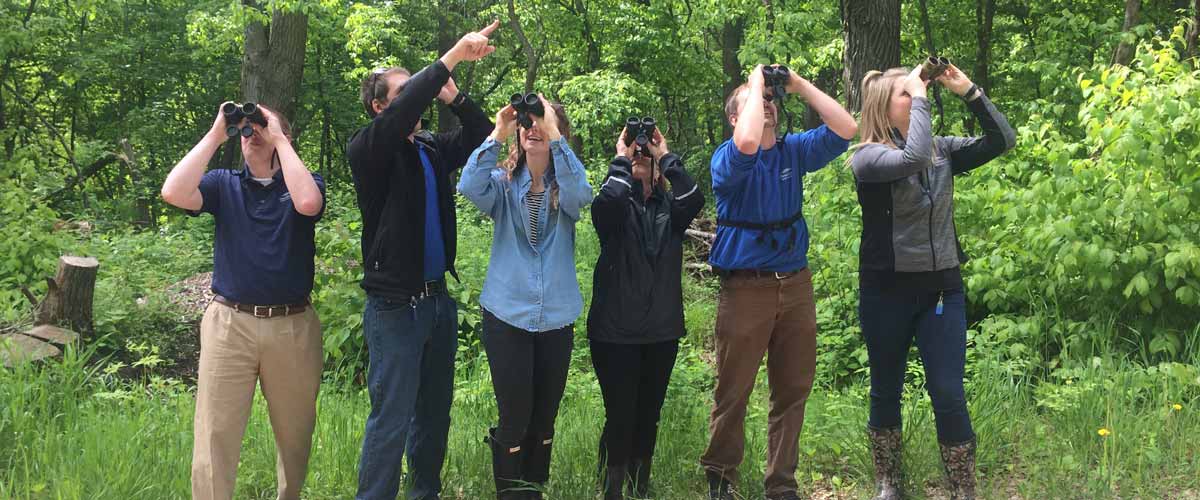 Six people look through binoculars in front of a green, wooded area.