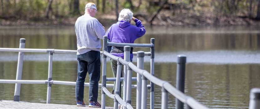 Two white-haired people stand by a rail and look across water.