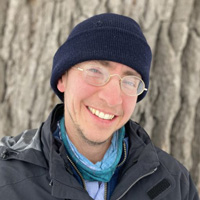 Alan, wearing a navy, knit beanie and wearing glasses, smiles in front of a tree.