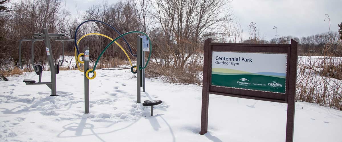 An outdoor gym surrounded by snow, with a sign in front that reads "Centennial Park Outdoor Gym" with the Three Rivers Park District logo and the city of Brooklyn Center logo.