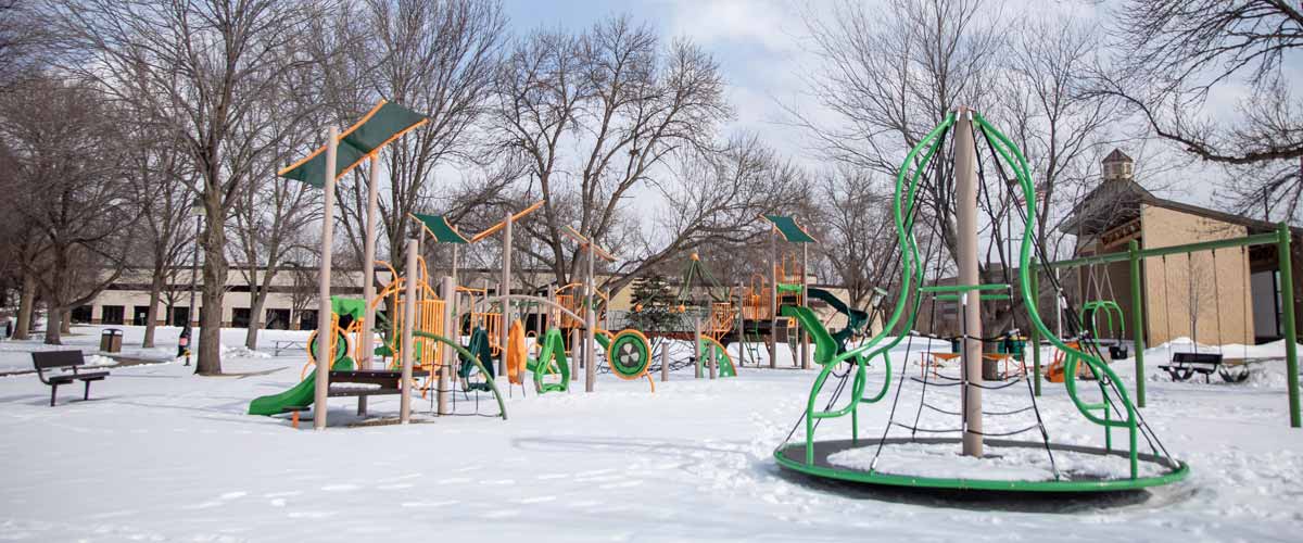 A green and yellow play area with a merry-go-round, swings, slides and other amenities, covered in snow.