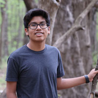 Alex, wearing glasses and a navy tee-shirt, stands in the forest in front of trees and branches.
