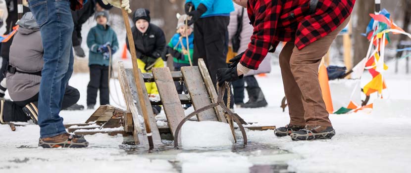 Two people grab move a block of ice onto a wooden ramp in a lake as people in the background look on.