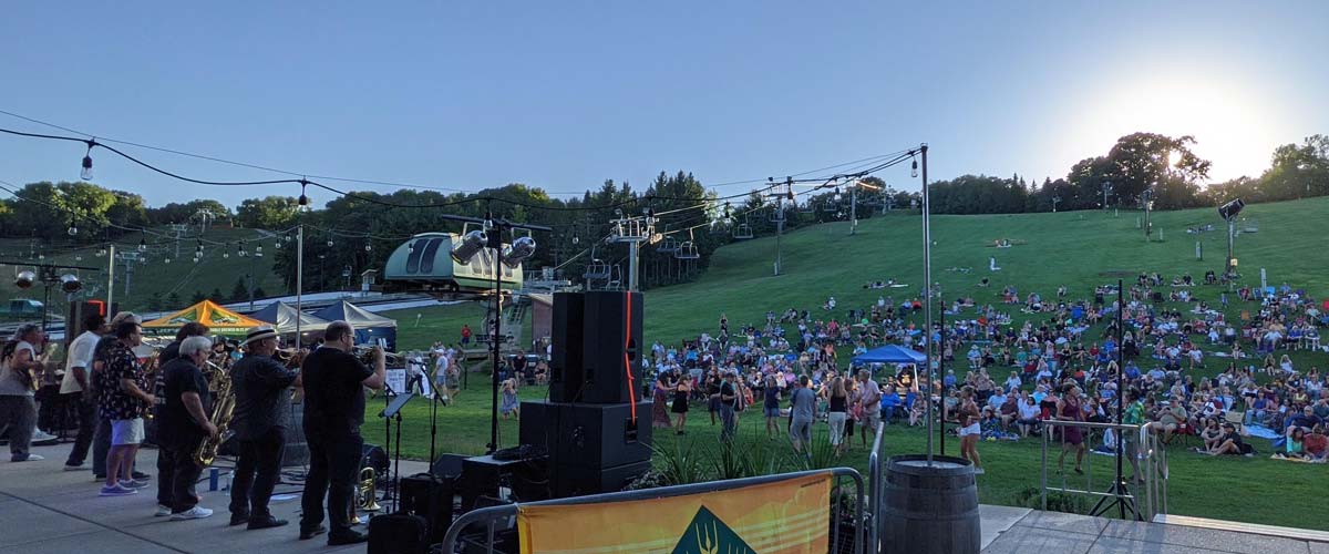 A crowd of people sit on the hill enjoying live music.