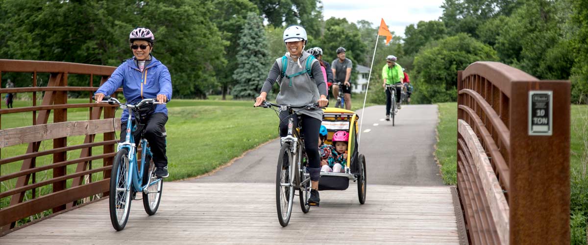 Photo of two adults biking across a bridge, one pulling children in a bike trailer behind their bike.