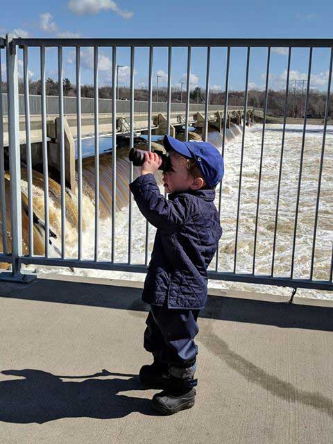 A boy wearing a baseball cap peers through binoculars, with a dam in the background.
