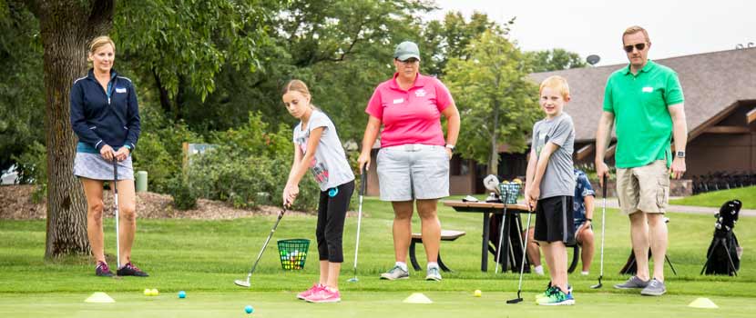 Three adults stand behind two kids as they learn how to golf.