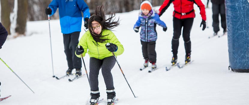 A girl in a yellow coat cross-country skis down a small hill while others follow behind.