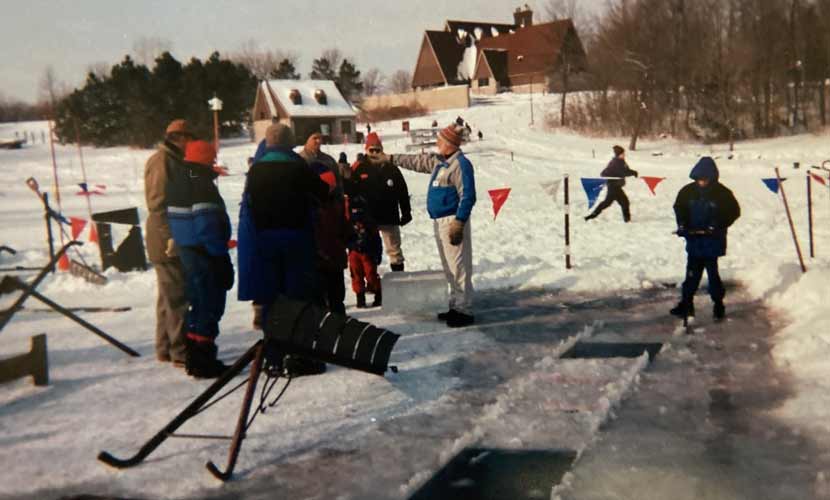 A group of people stand on a frozen lake, near a strip of ice that has been cut into blocks, at the 2000 Hyland ice harvest event.