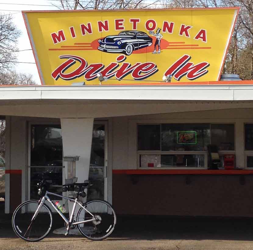 a white bike parked in front of a drive in with a large yellow sign on top that says Minnetonka Drive in.