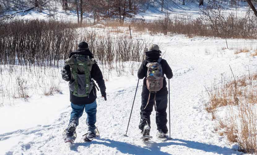 Two people dressed in winter gear with backpacks snowshoe down a trail.