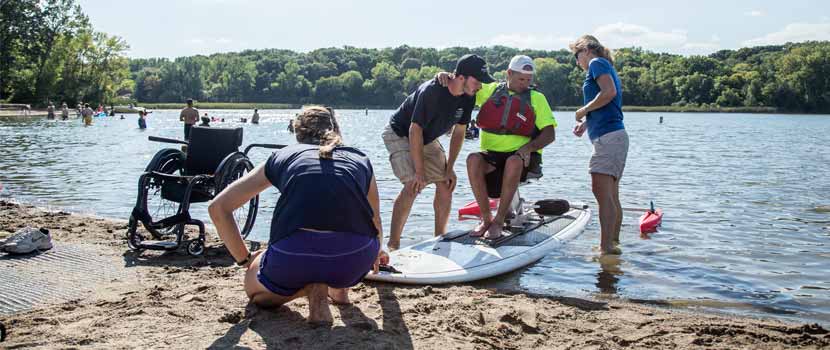 three rivers staff help a man onto an adapted paddleboard. his wheelchair is in the sand next to them.