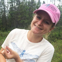 a woman in a white shirt and pink ball cap smiling while holding a dragonfly.