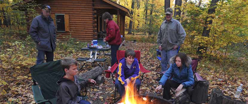 a family sitting around a campfire in the fall in front of a log cabin.