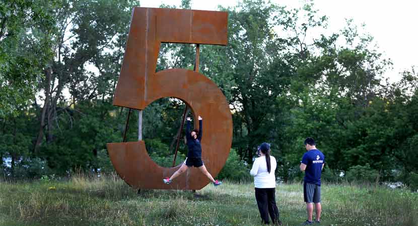 a person jumps in front a metal 5 sculpture while two others take her photo.