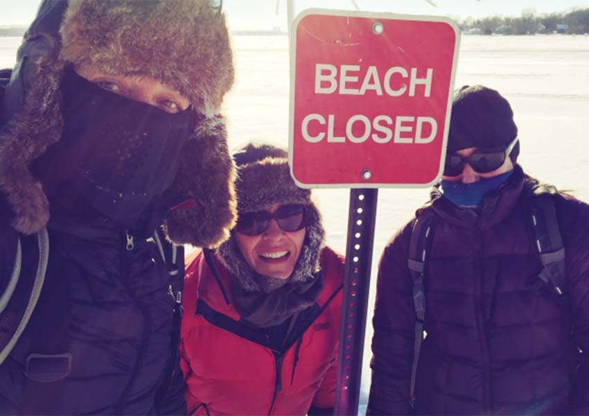 Three people in winter hats and coats stand around a red sign that says "beach closed."