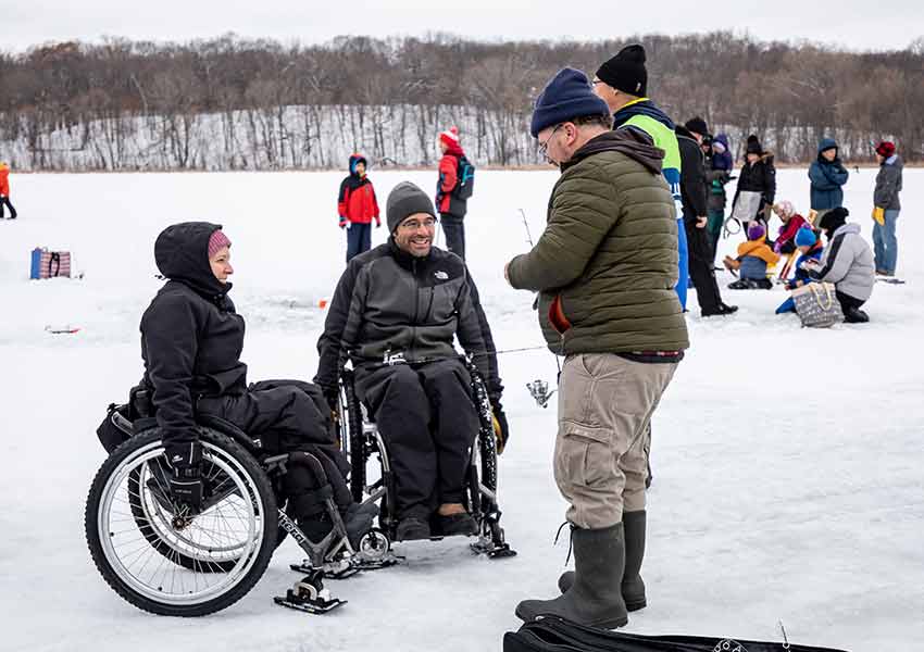 a man and a woman dressed in full winter layers in wheelchairs next to a man standing ice fishing on a frozen lake