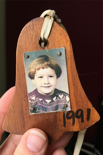 a wooden bell ornament with a photo of a young girl with a bowl cut pasted onto it. The corner says 1991 in black marker.